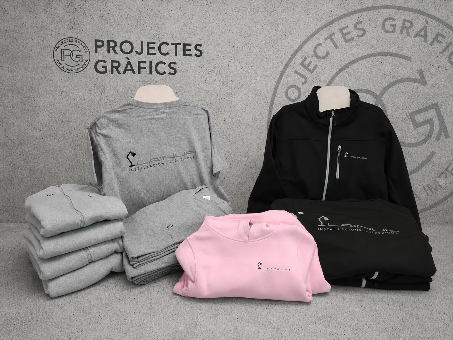 Ropa laboral personalizada con camisetas, sudaderas y chaqueta corporativa sobre fondo de Projectes Gràfics.
